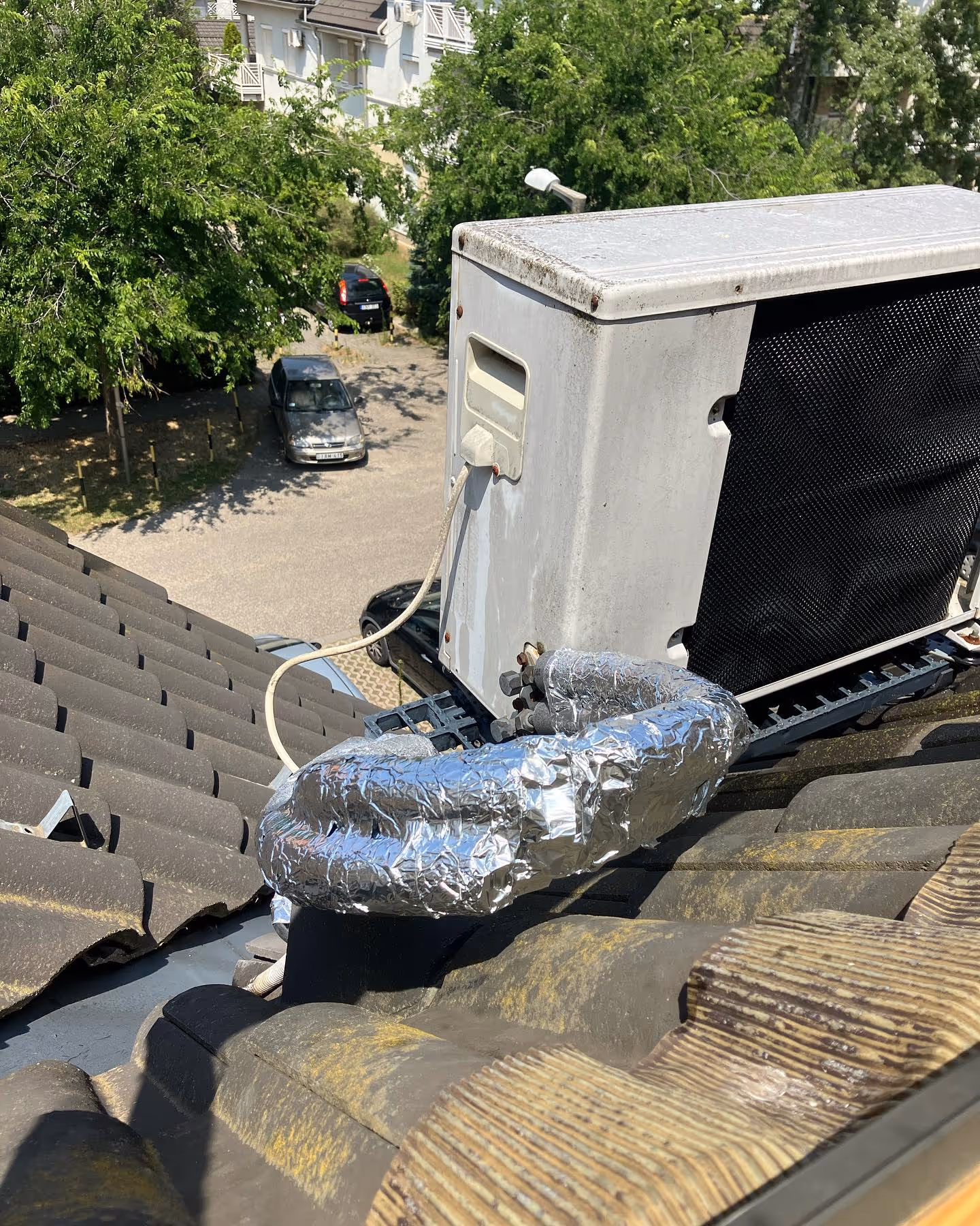 Air conditioning unit installed on a tiled roof with insulated pipes wrapped in silver foil.