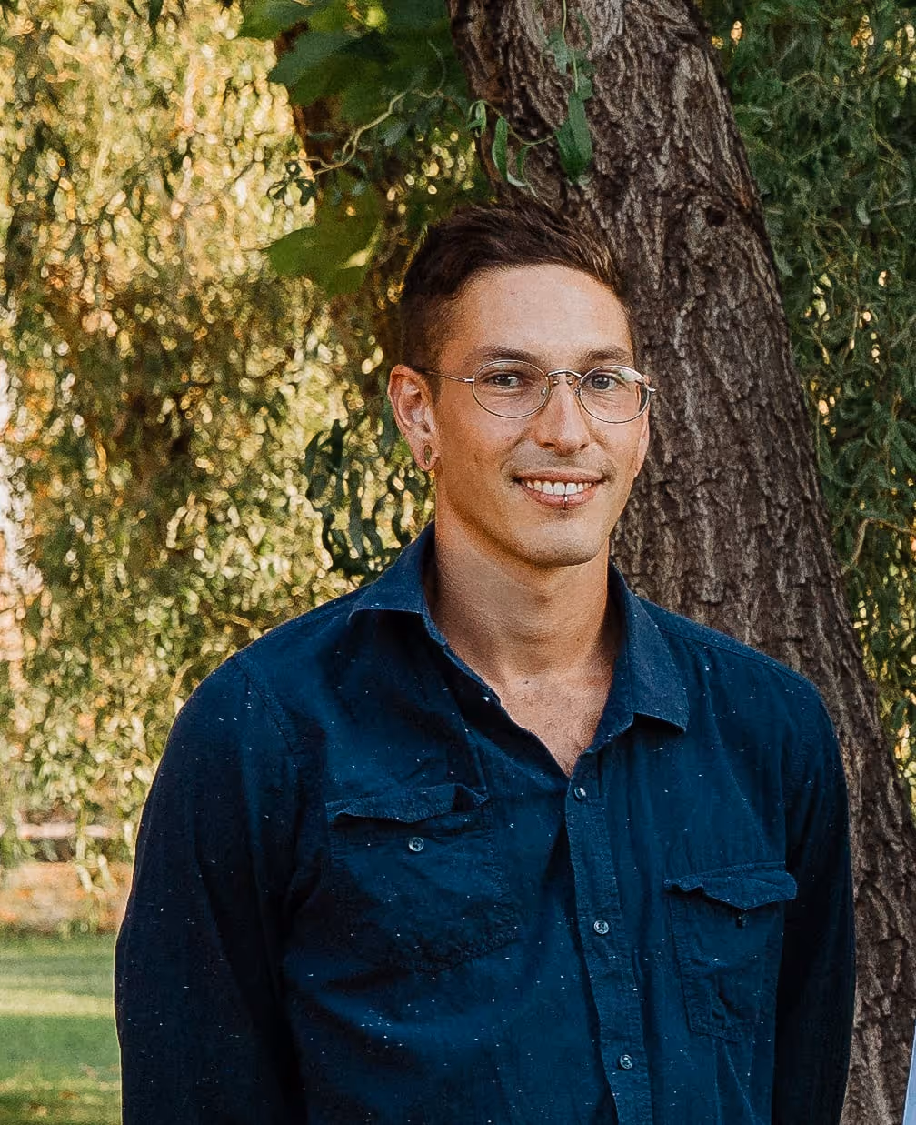 Smiling man with glasses and short dark hair wearing a blue shirt standing outdoors near a tree.
